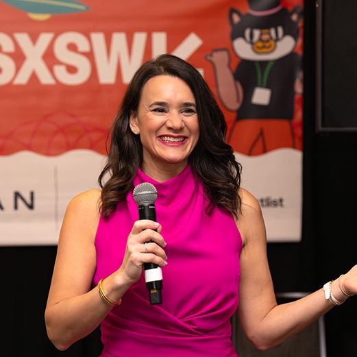 Woman speaker gestures on stage at SXSW conference with colorful branded backdrop and audience.