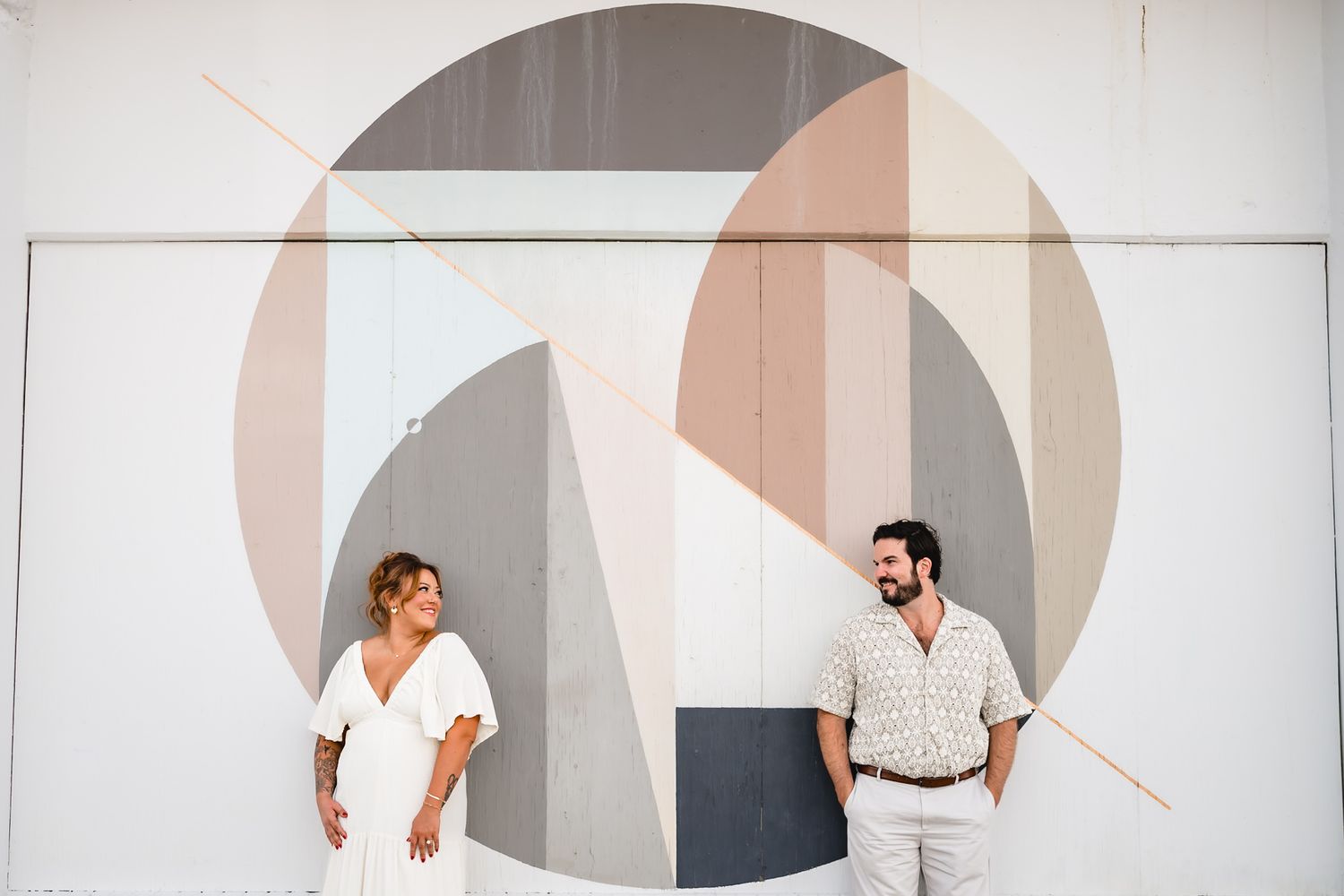 Couple smiling at each other standing in front of a colorful geometric mural outdoors.