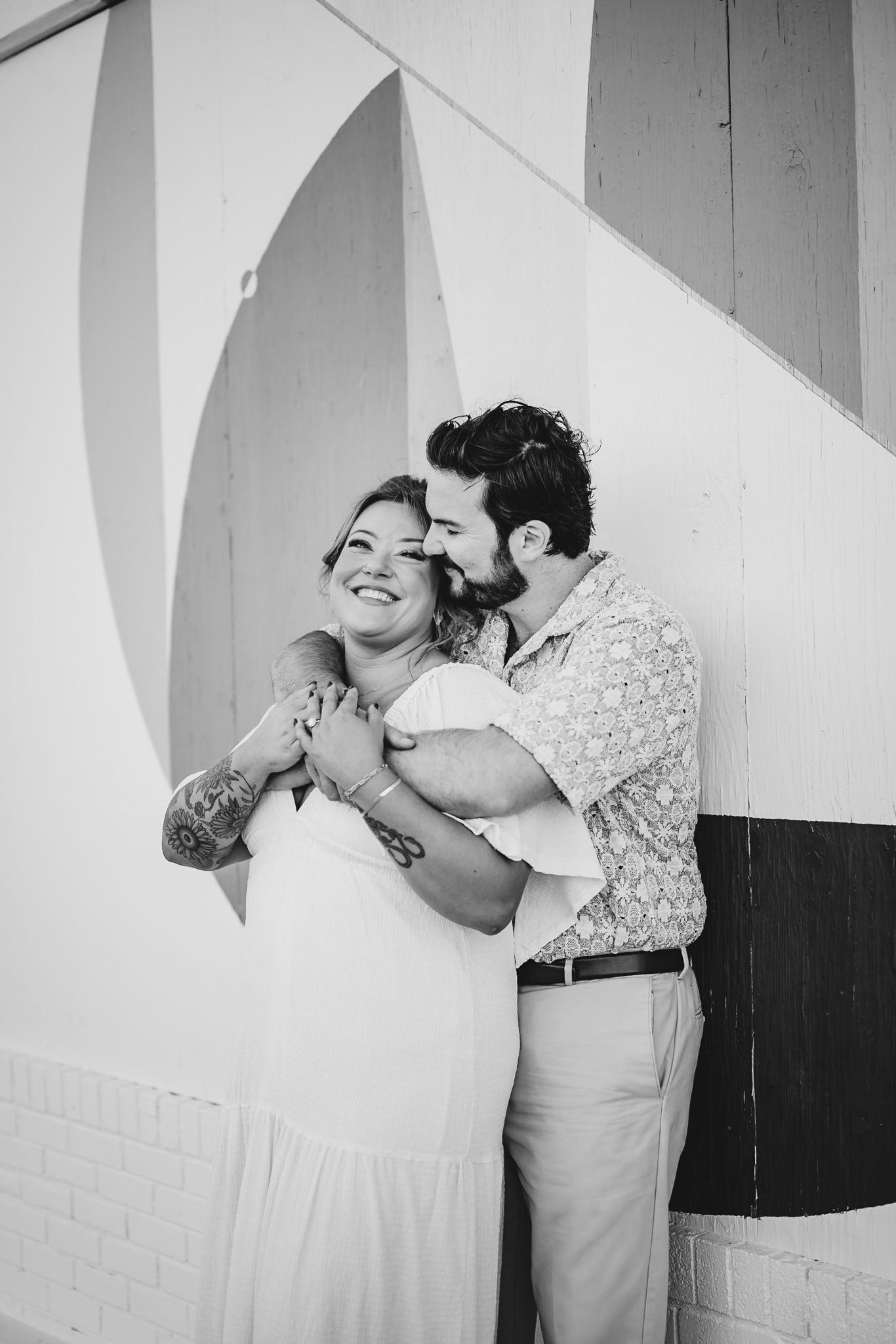 Black and white photo of a couple embracing, man kissing woman's cheek, she smiles, tattoos visible, against a painted wall.