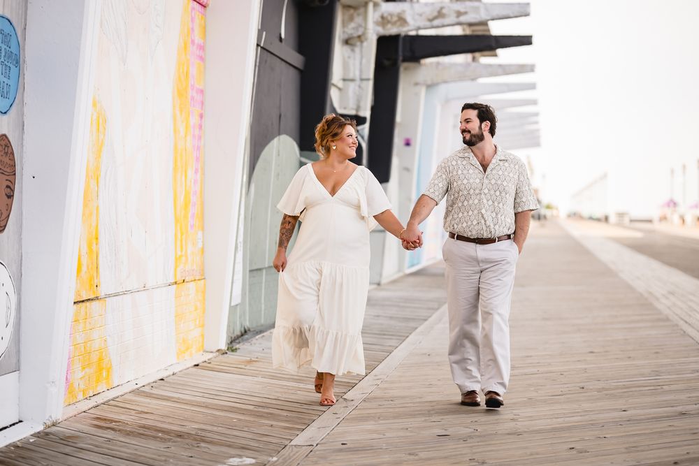 A couple walks hand-in-hand on a boardwalk, dressed in white and beige outfits, with colorful murals in the background.