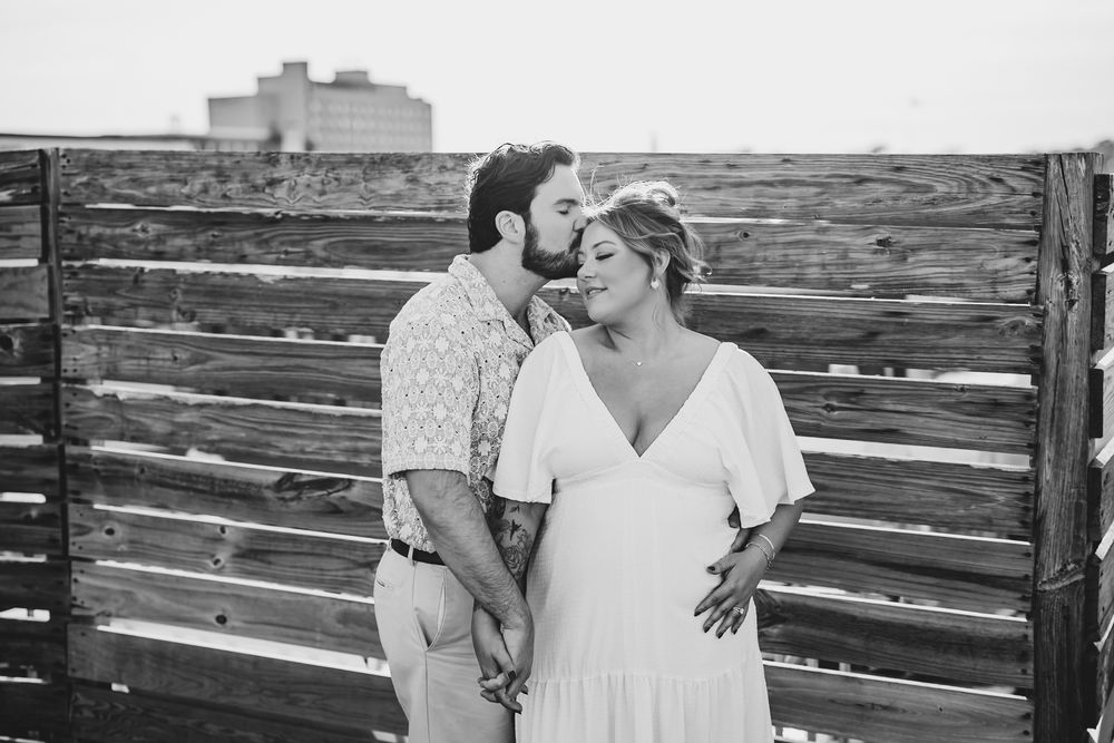 Black and white photo of a couple holding hands, man kissing woman's cheek near a wooden fence outdoors.