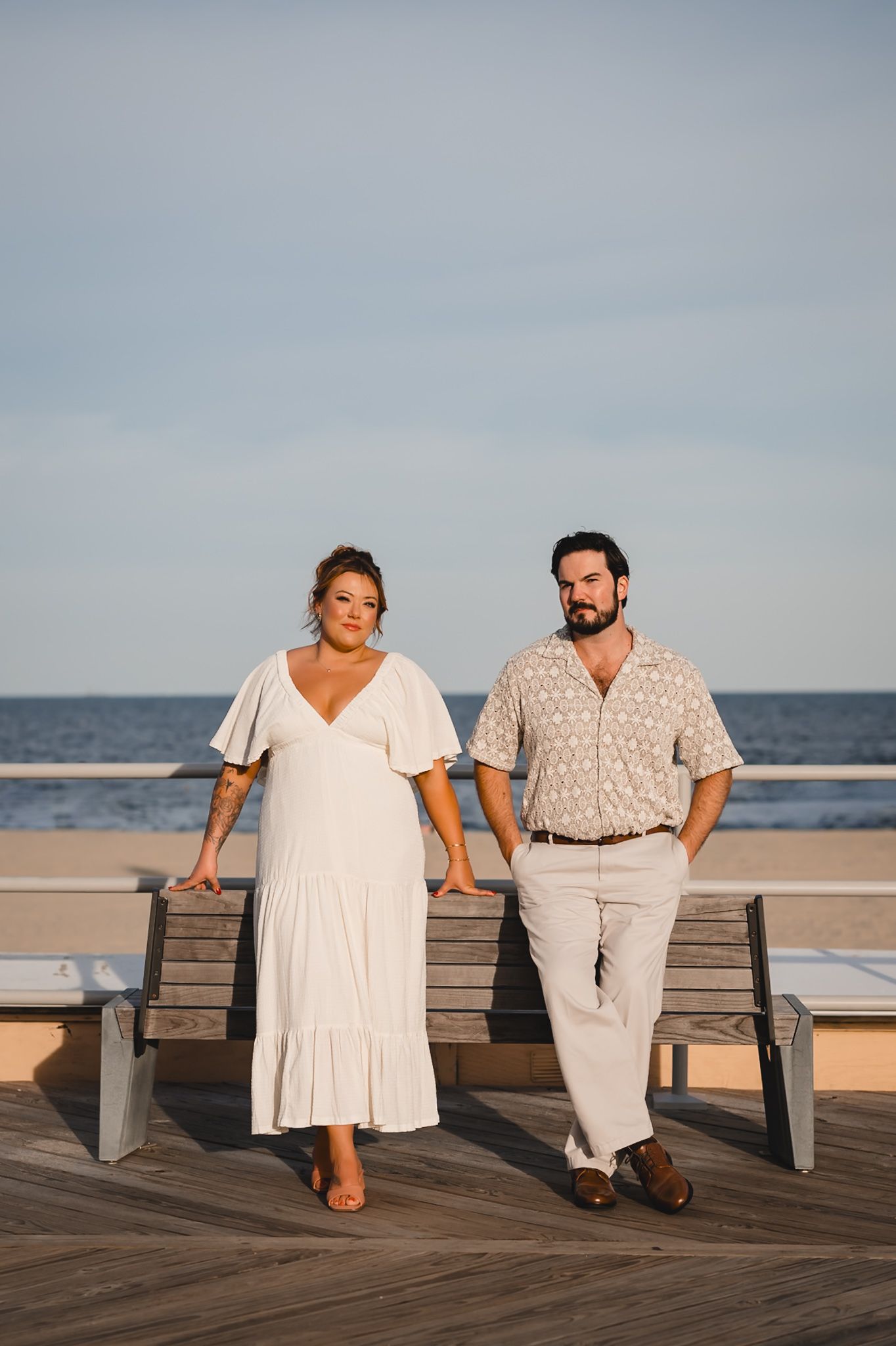Couple posing on a boardwalk by the ocean, woman in white dress, man in beige shirt and pants.