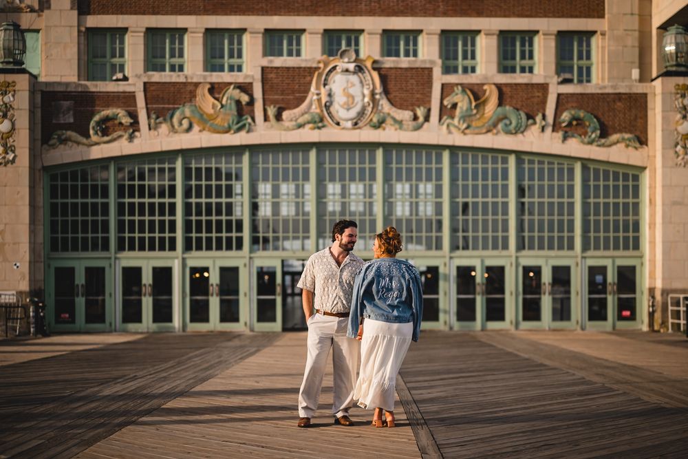 Couple holding hands on boardwalk in front of ornate historic building with large arched windows at golden hour.