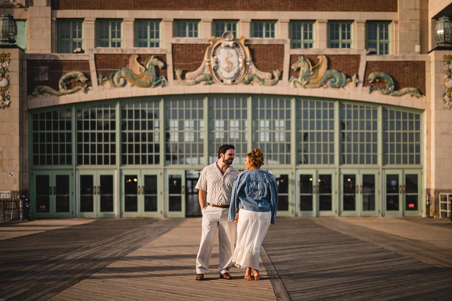 Couple holding hands on boardwalk in front of ornate historic building with large arched windows at golden hour.
