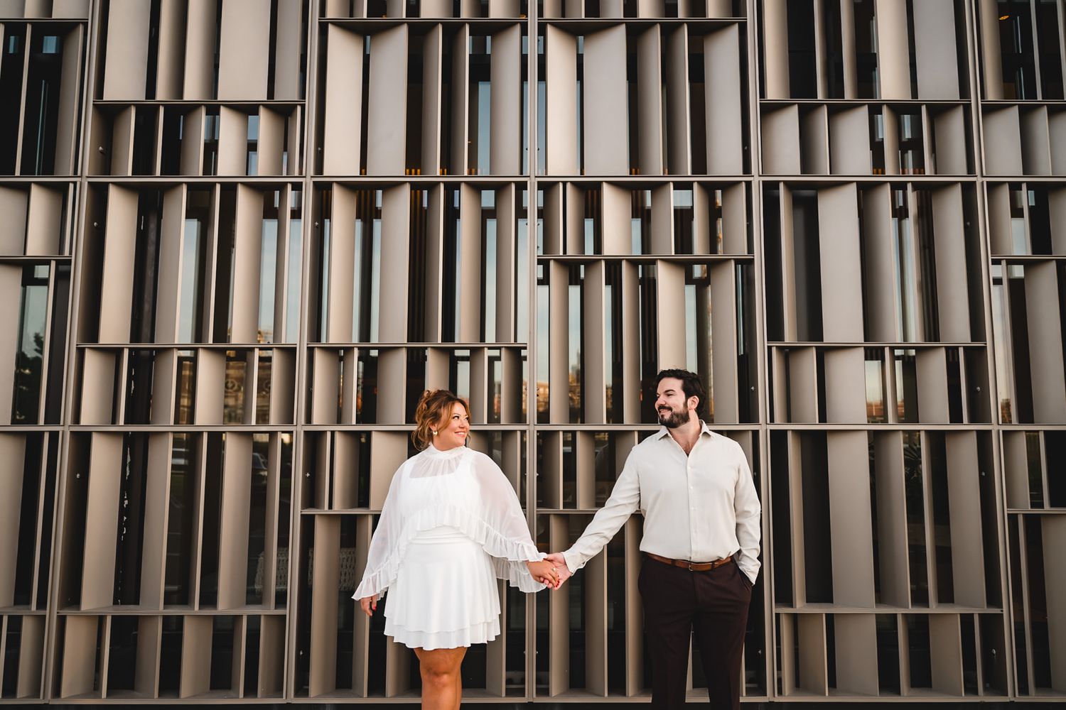 Couple holding hands in white outfits standing before a modern geometric metal wall facade.