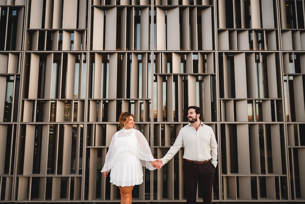 Couple holding hands in white outfits standing before a modern geometric metal wall facade.