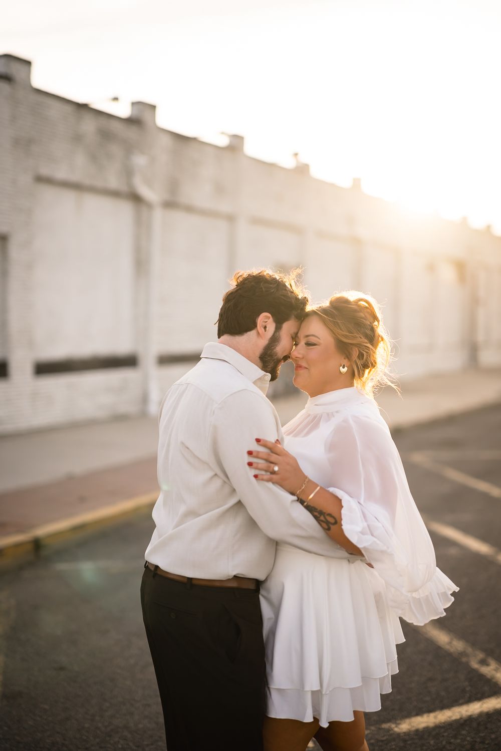 Couple embracing romantically at golden hour sunset in a parking lot, both dressed in white outfits.