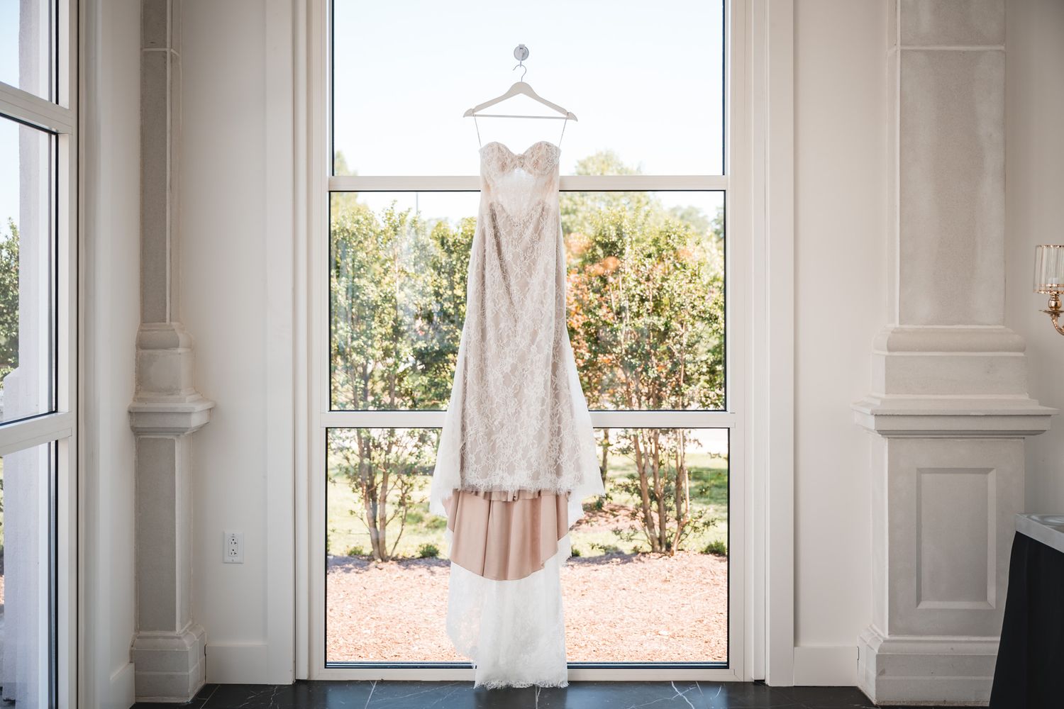 Elegant strapless wedding dress hanging in a sunlit window with white walls and garden views.