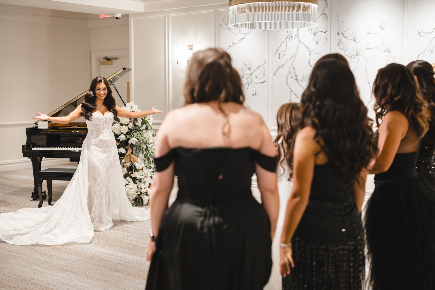 Bride in white gown stands near grand piano gesturing to bridesmaids in black dresses in elegant wedding venue.