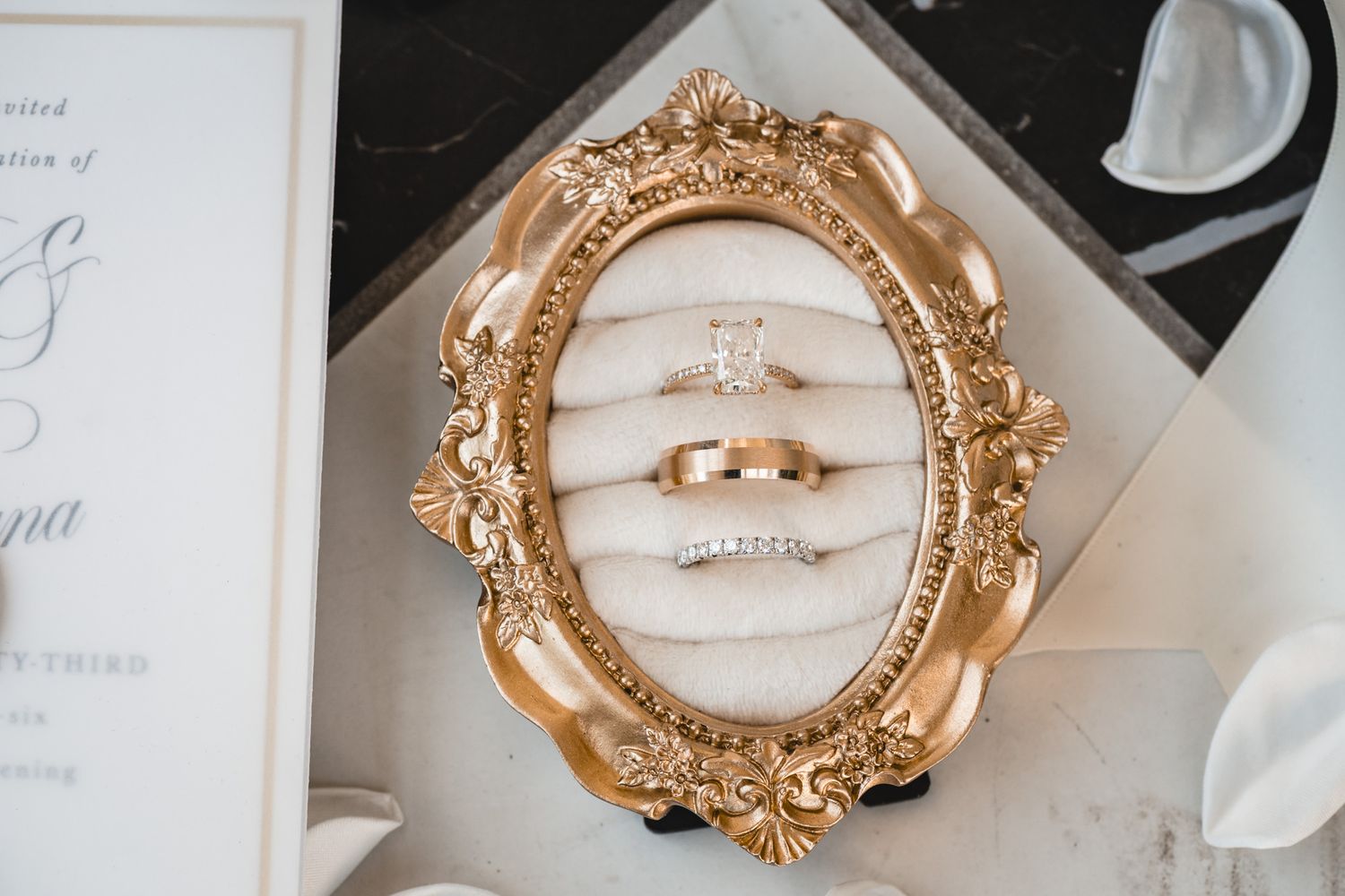 Gold ornate frame displaying an engagement ring, wedding band, and diamond eternity band on ivory velvet.