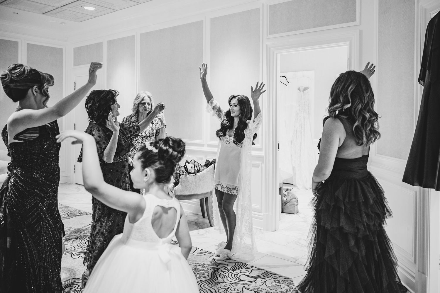 Bridesmaids celebrating with the bride in a hotel room, raising arms in joy, black and white wedding photo.
