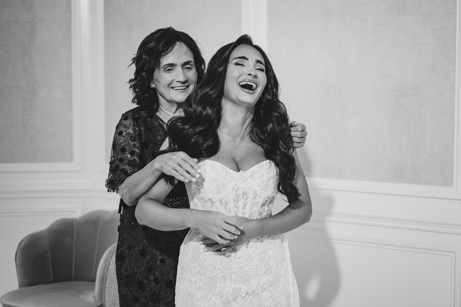 Black and white photo of a laughing bride in a lace gown embracing an older woman at a wedding.