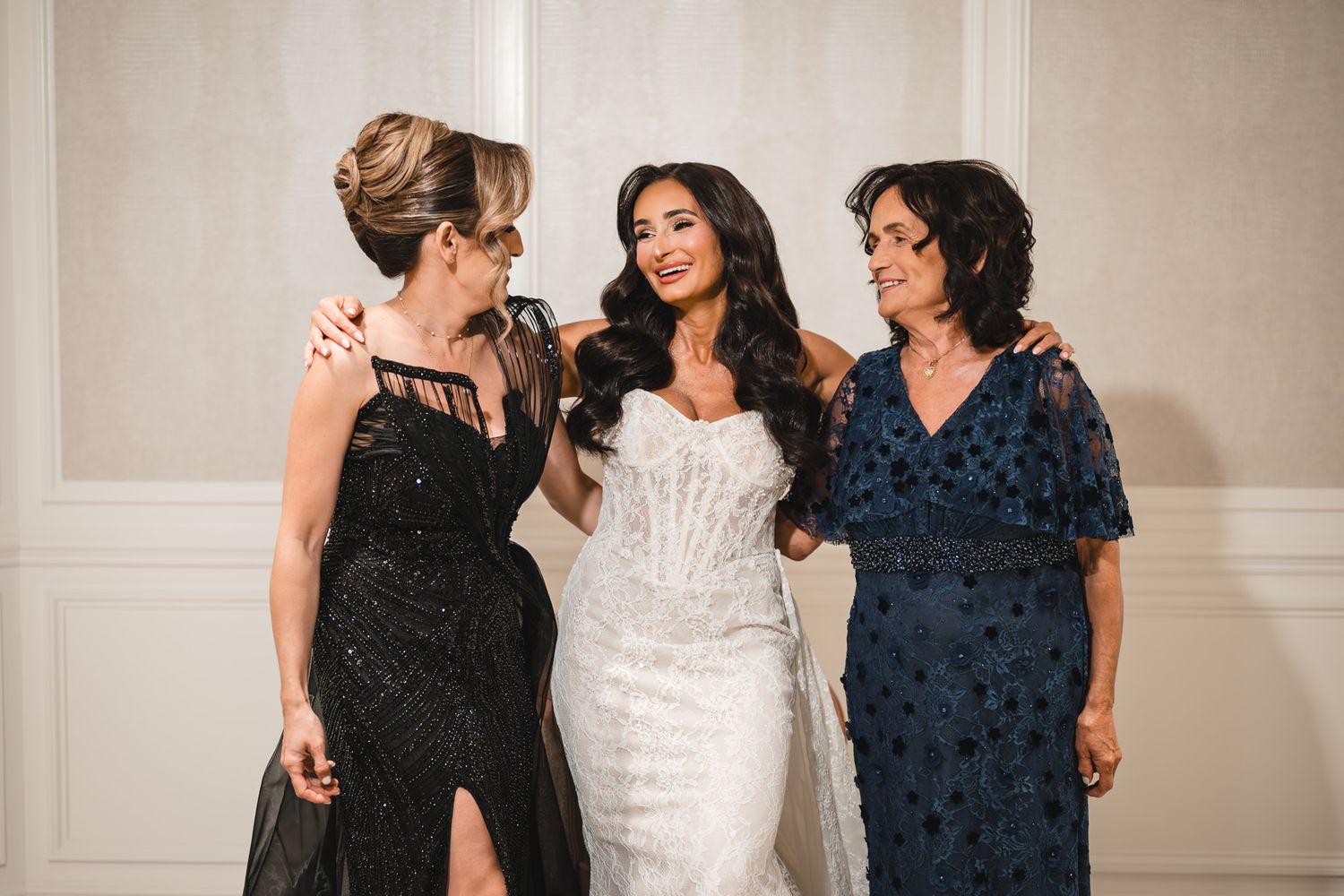 Bride in white corset gown poses with two women in elegant black and blue dresses at a formal wedding celebration.