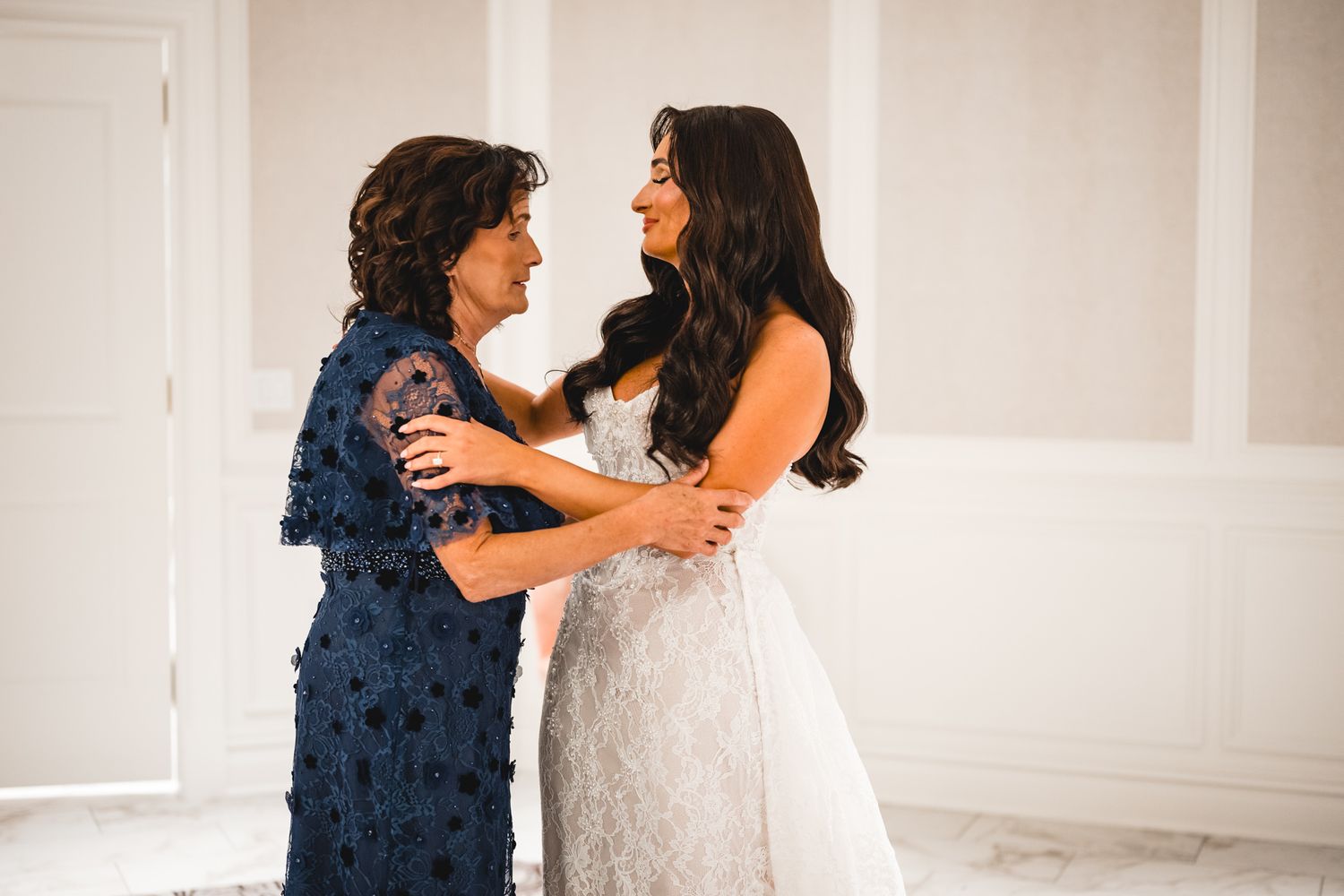 Bride in lace gown shares a joyful embrace with her mother in a navy blue floral dress.