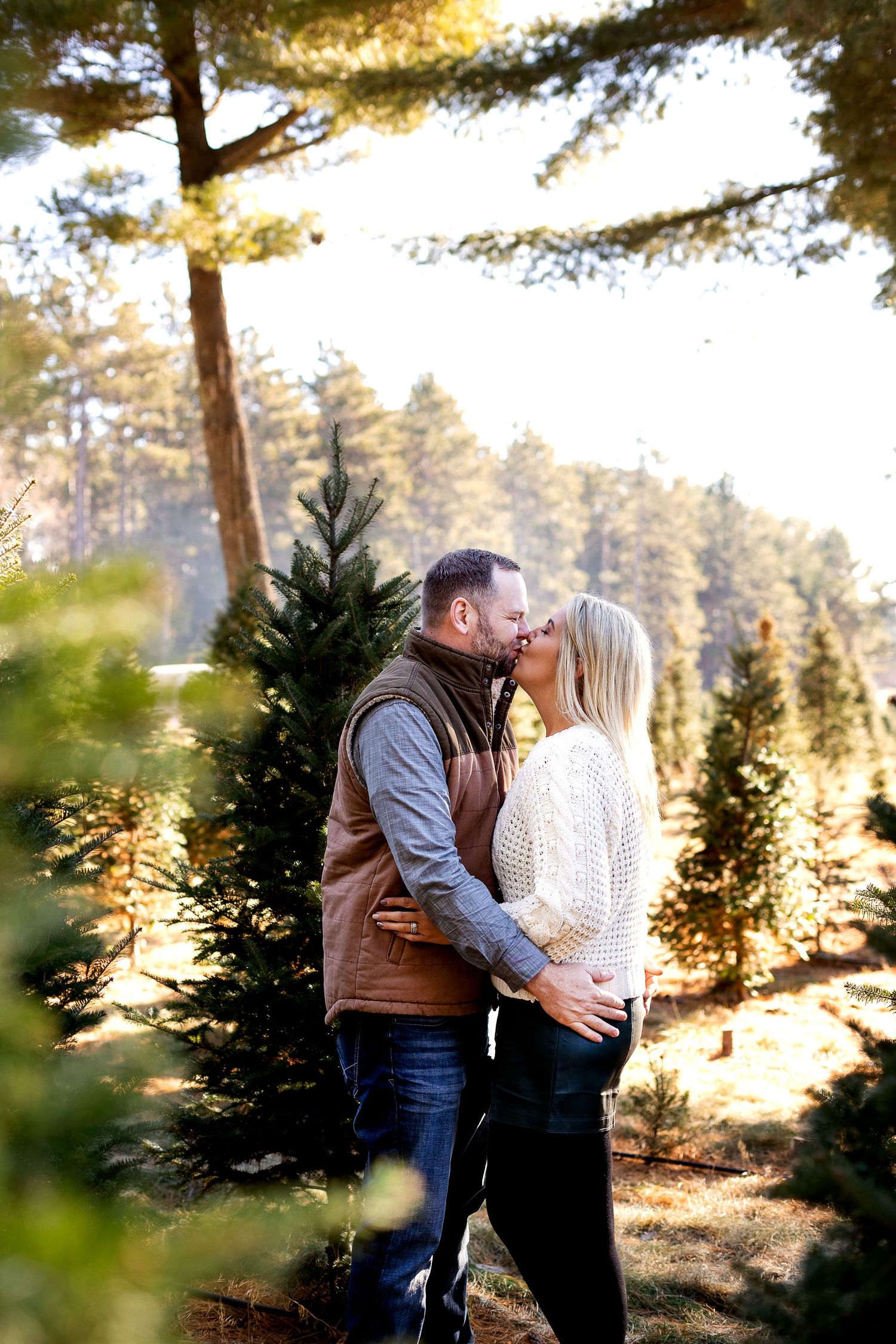 Couple kissing among evergreen trees at a Christmas tree farm, woman in white sweater, man in brown vest, sunny forest backdrop.
