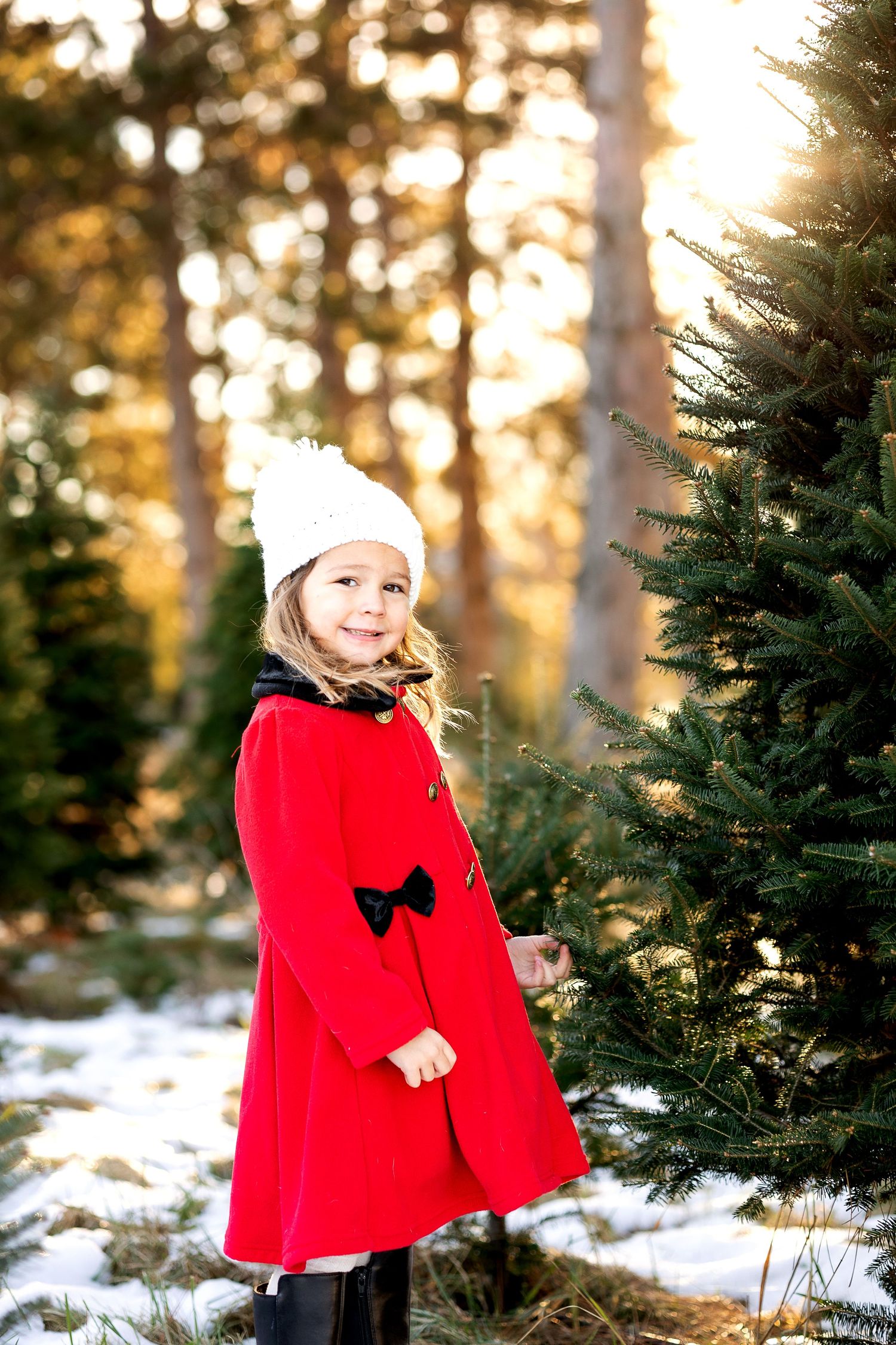 Young girl in red coat and white knit hat smiling beside a Christmas tree in a snowy forest.