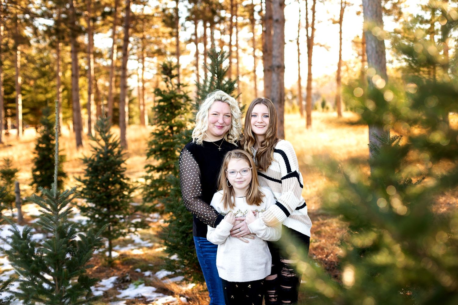 Three generations of women smiling together among pine trees in a snowy forest during golden hour.