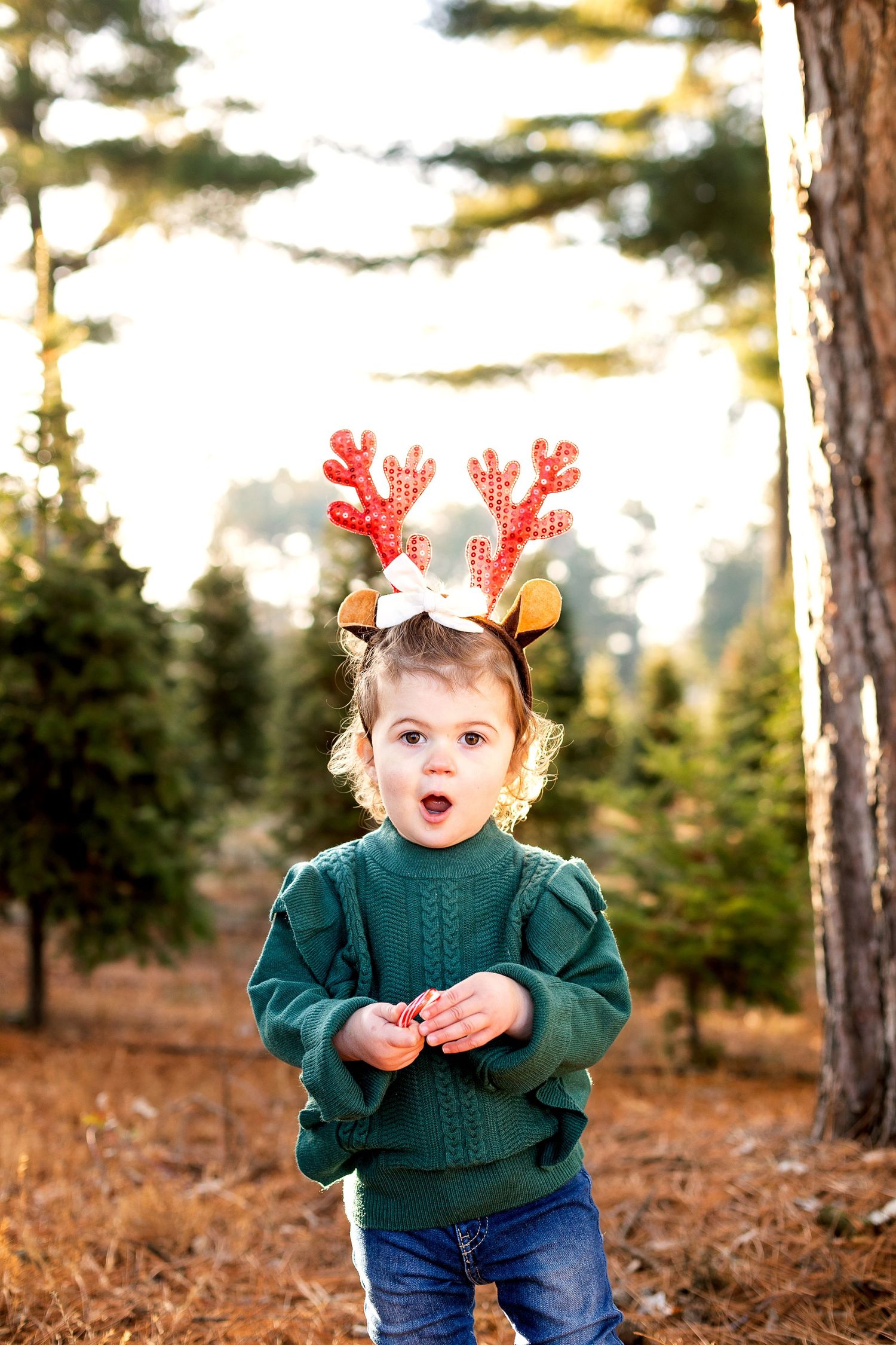 Toddler girl in green sweater and reindeer antler headband standing among Christmas trees at a tree farm.