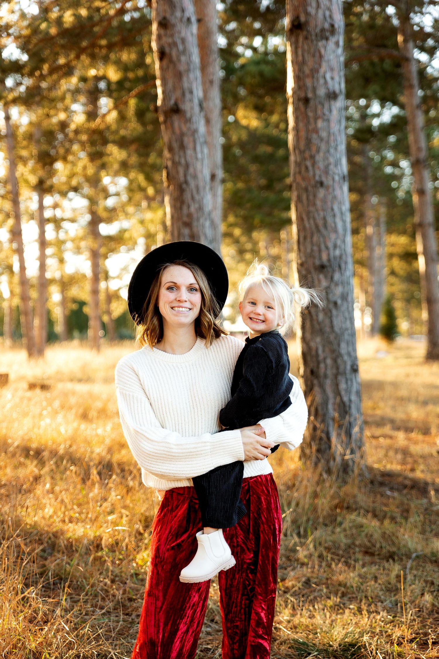 A smiling woman in a white sweater and red velvet pants holds a young blonde child in a sunlit pine forest.
