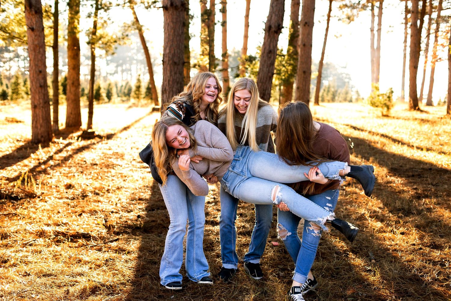 Four girls laughing and playing in a sunlit forest, two carrying friends on their backs among tall pine trees.