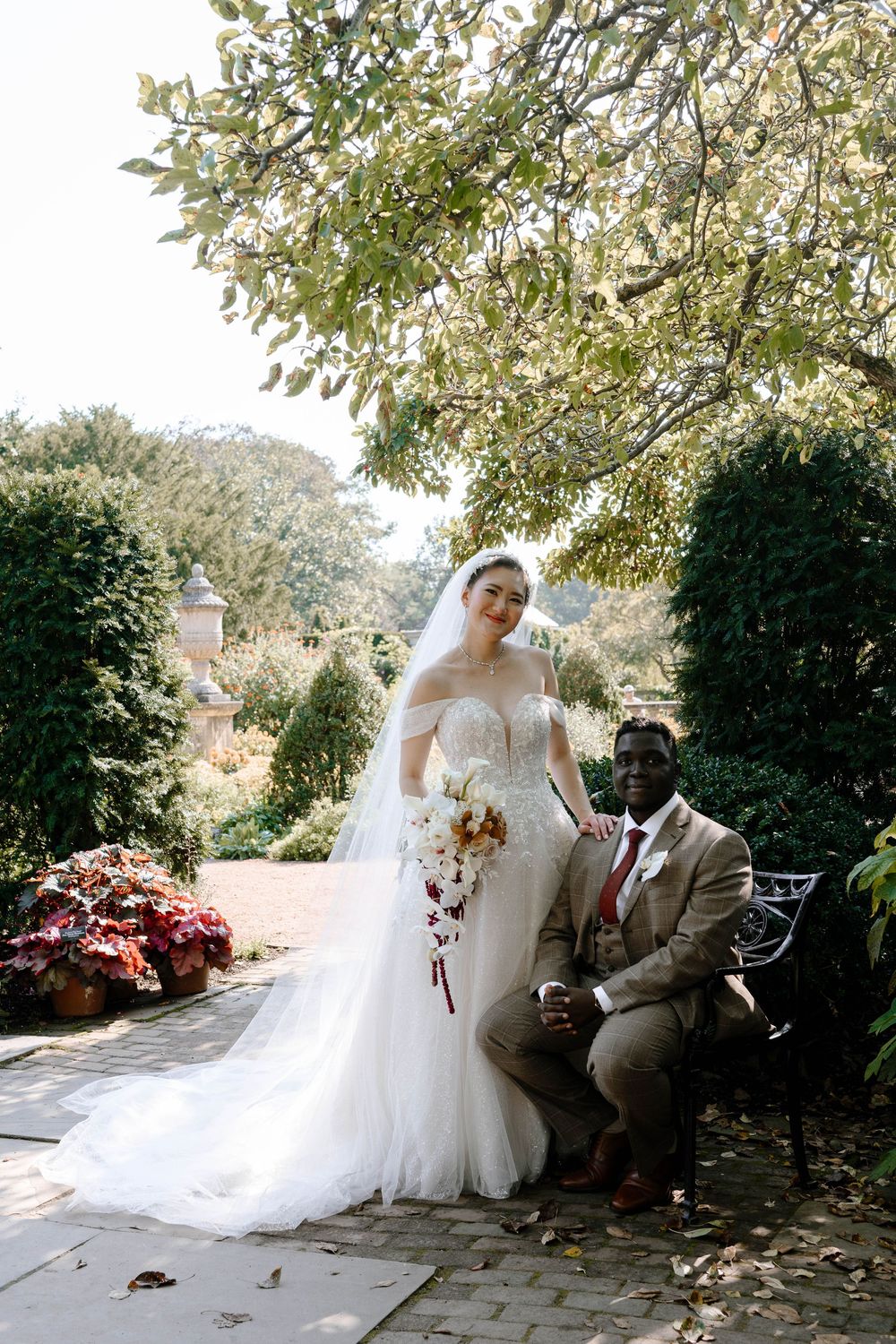 Bride in white gown with veil holds bouquet beside groom in tan suit seated outdoors in garden setting.