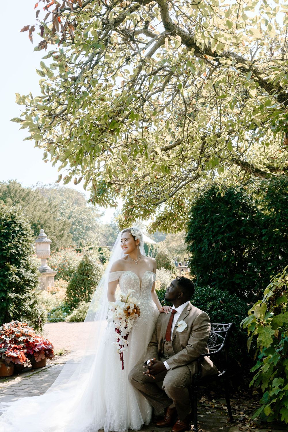 Interracial wedding couple posing under autumn tree, bride in white gown with veil.