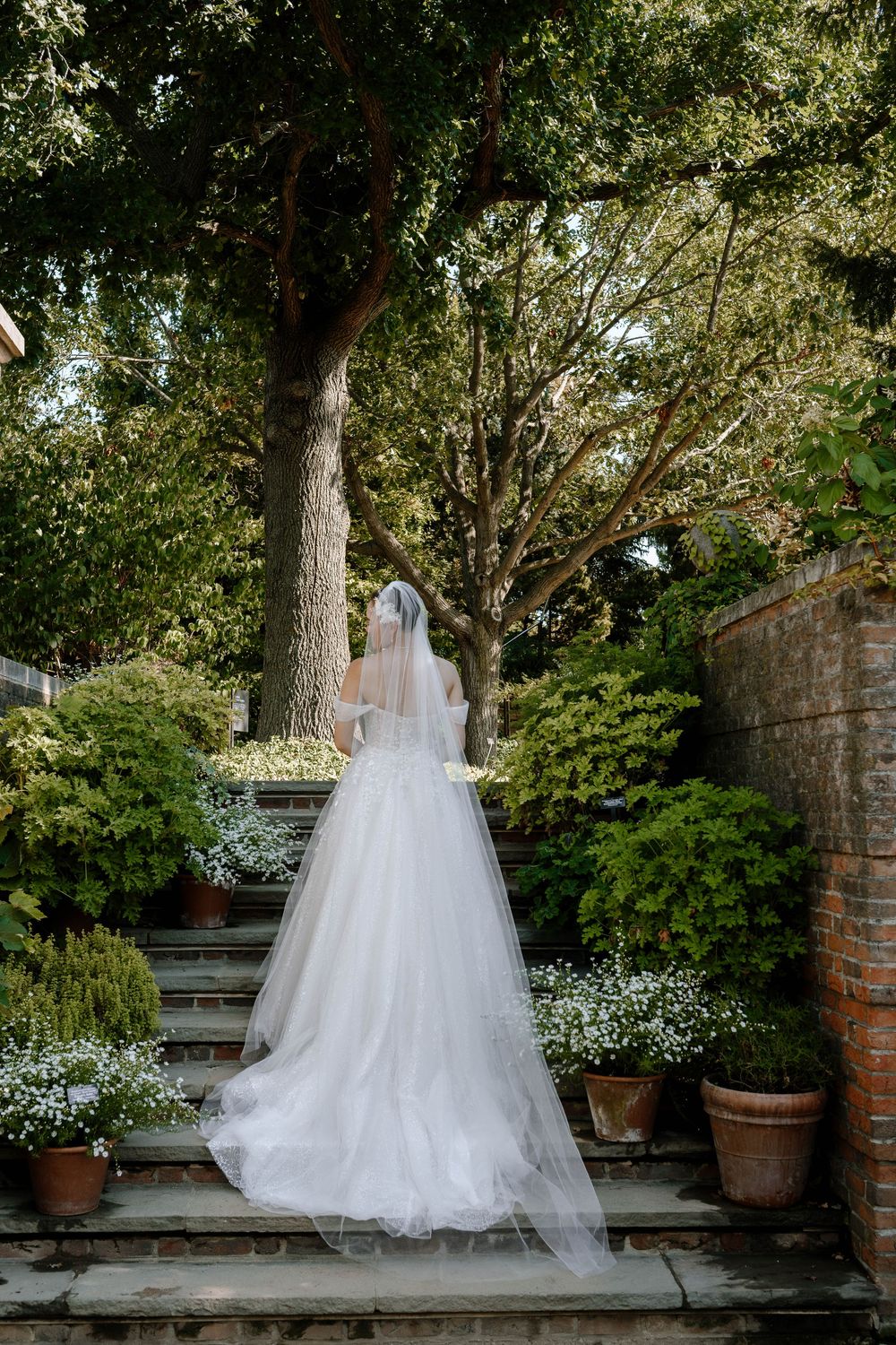 Bride in white gown and veil ascending garden steps surrounded by lush greenery and potted flowers.