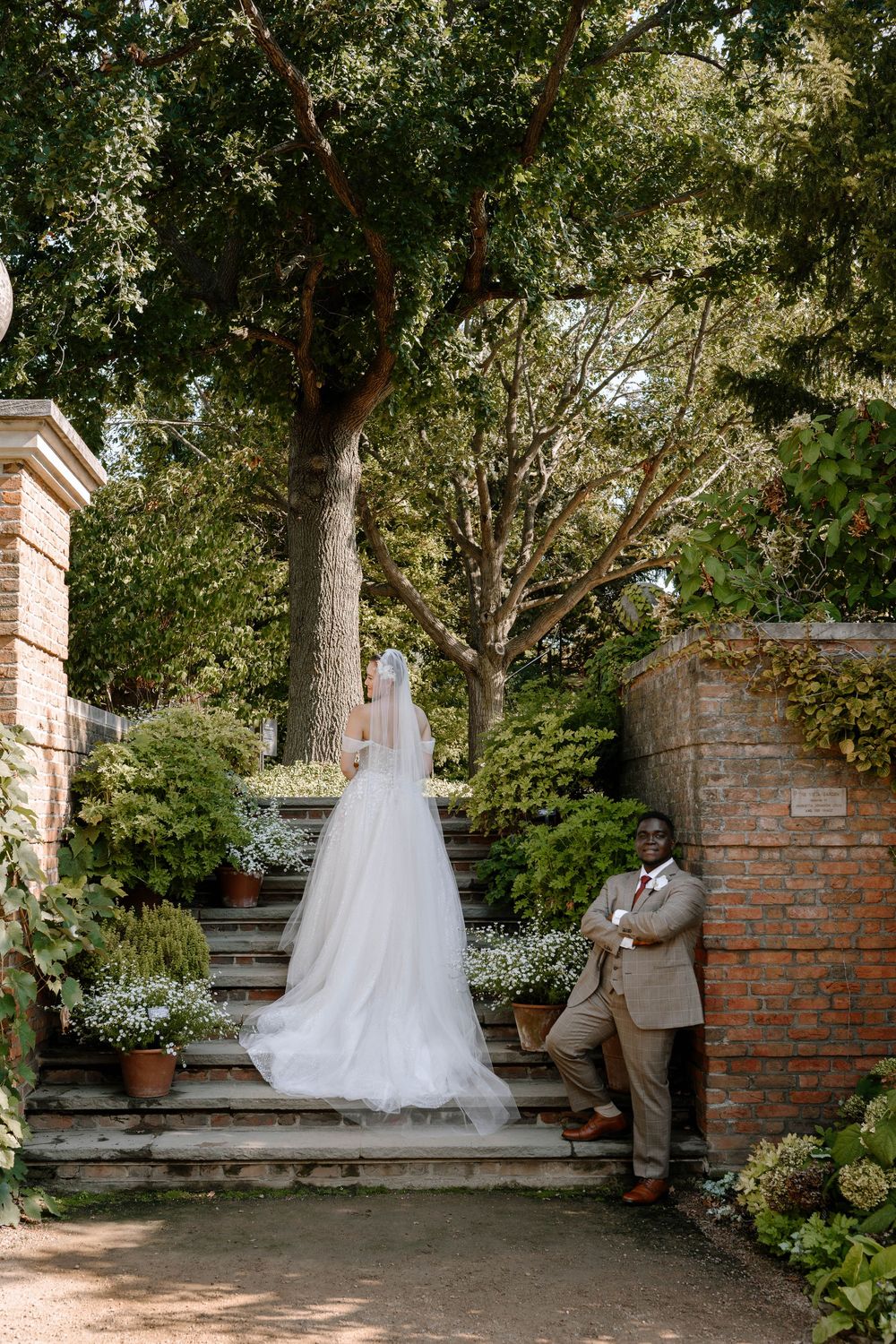 Bride in white gown and veil stands on garden steps as groom in tan suit leans against brick wall.