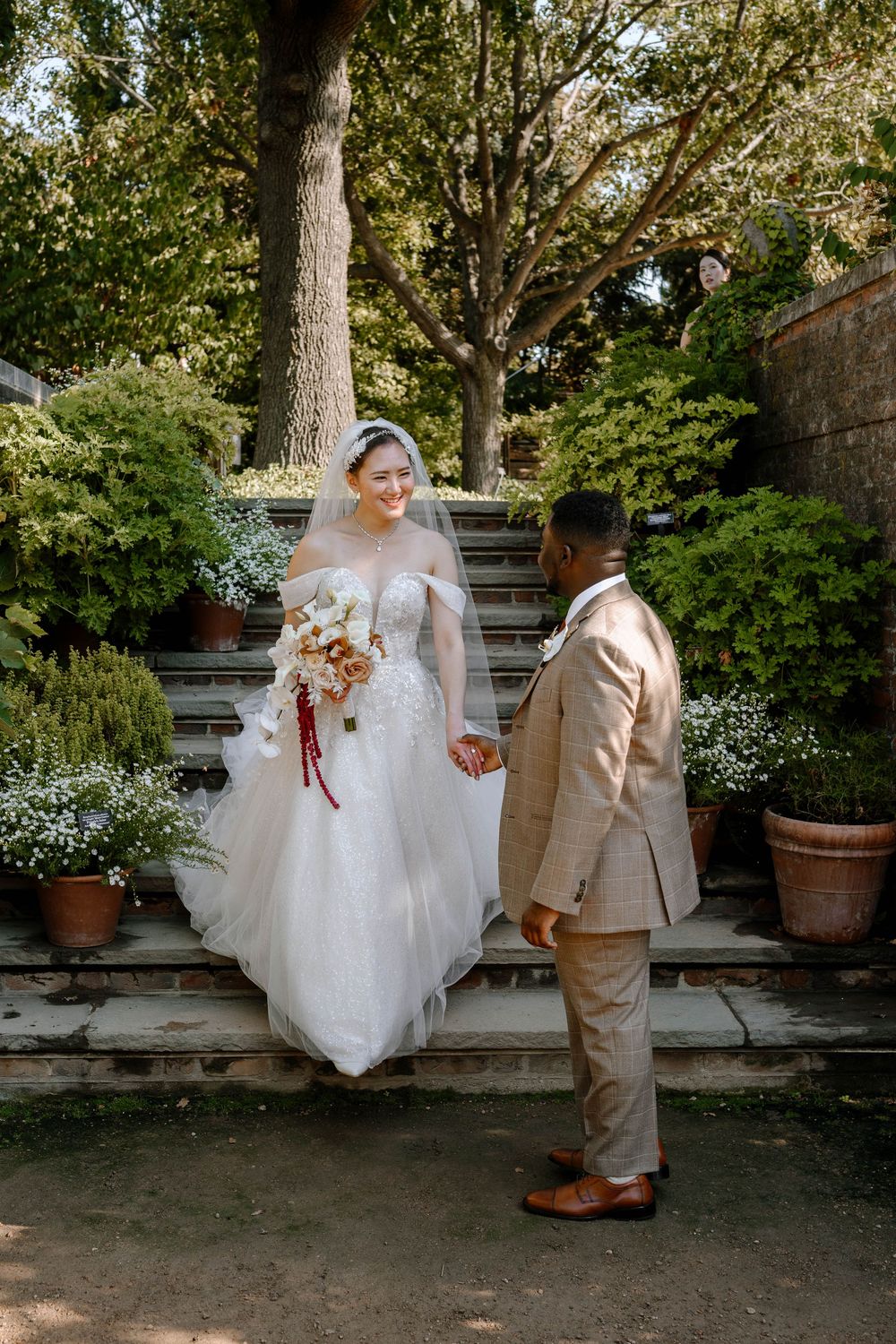 Bride and groom holding hands on garden steps, bride in lace gown with bouquet.