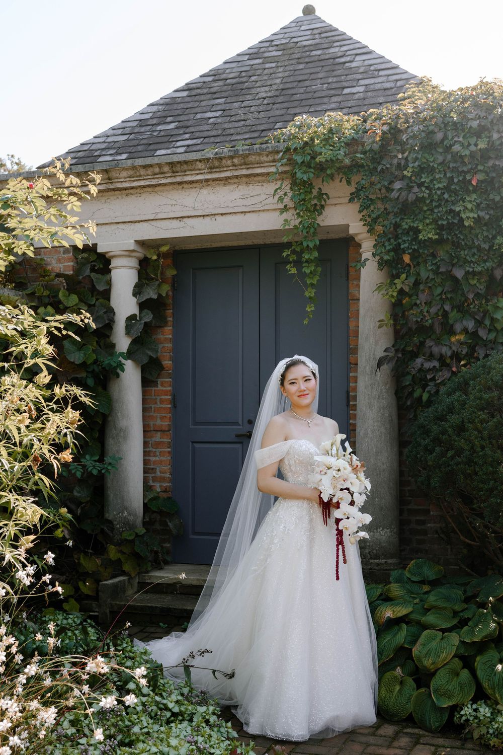 Bride in white off-shoulder gown and veil holds bouquet before ivy-covered brick building with gray door.