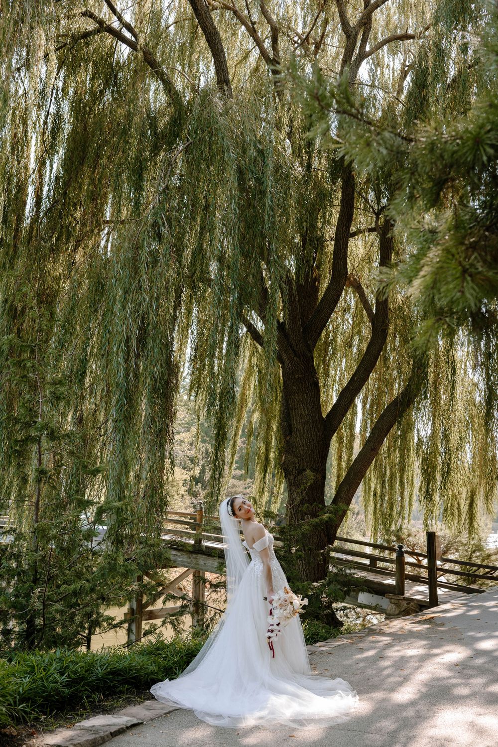 Bride in white gown and veil poses under weeping willow tree holding bouquet in lush garden setting.