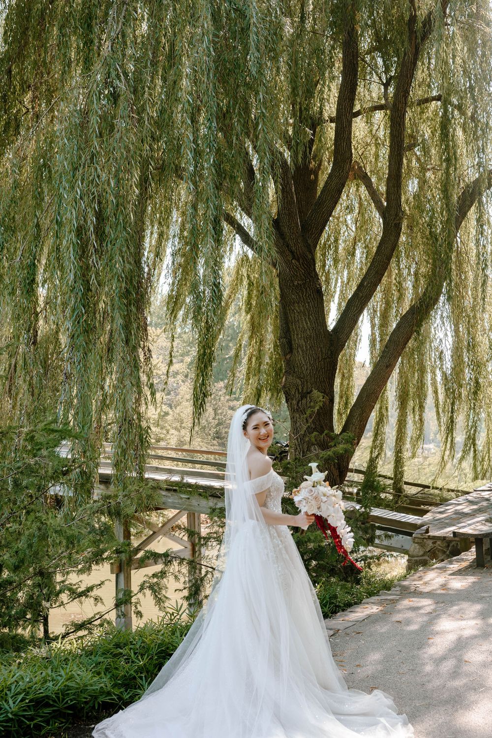 Bride in white gown and veil holds bouquet under a weeping willow tree on a garden path.