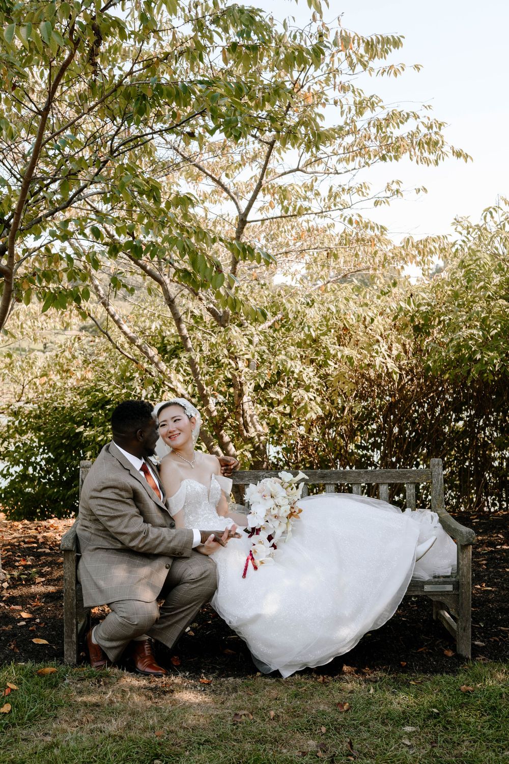 Bride and groom sitting on a wooden bench in a lush garden, sharing a joyful moment on their wedding day.