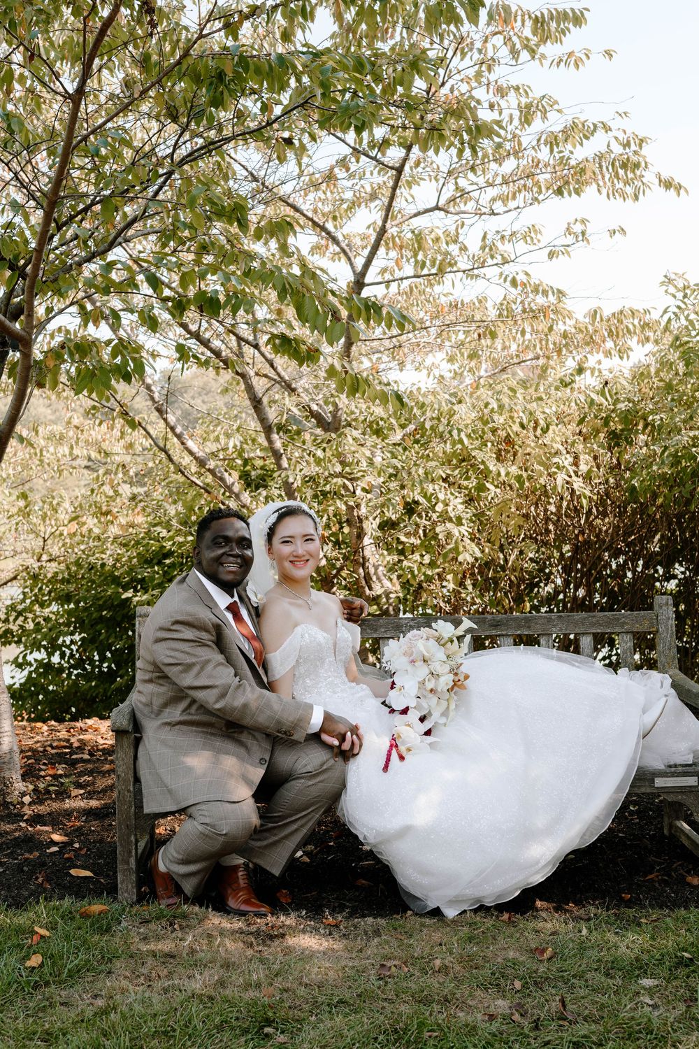 Smiling interracial couple in wedding attire pose together on a wooden bench surrounded by lush greenery.