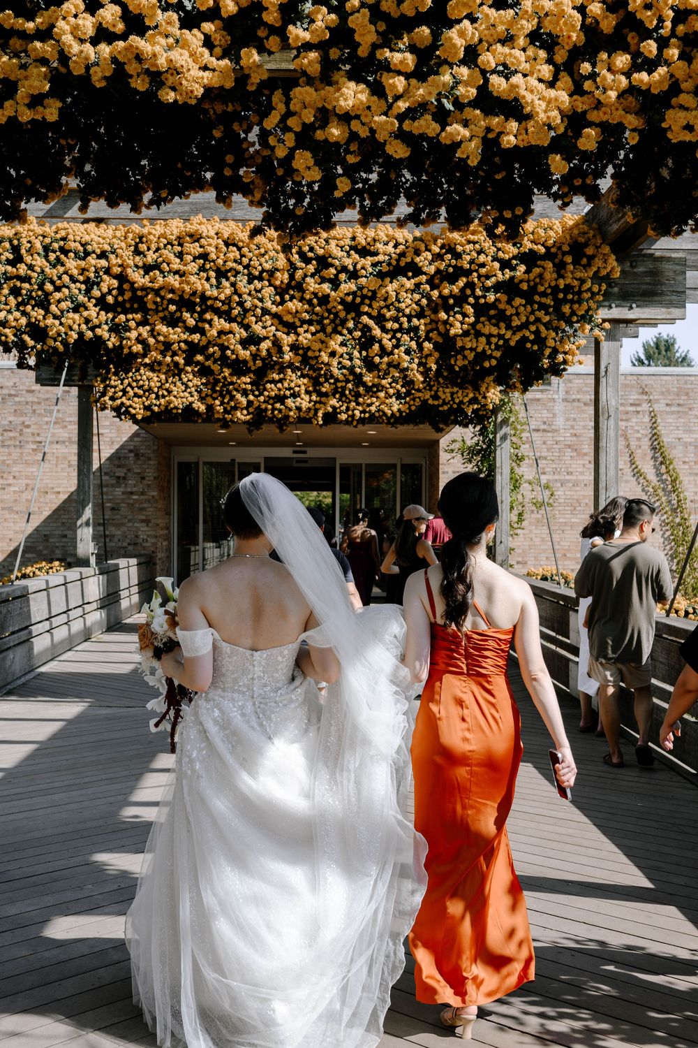 Bride in white gown and veil walks with bridesmaid in orange dress under yellow floral arch.