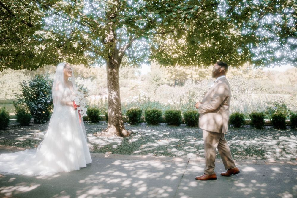 Bride in white gown and groom in tan suit share a first look moment under a lush green tree outdoors.
