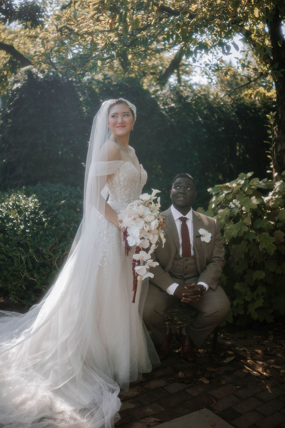 Bride in white gown with veil holds bouquet as groom in tan suit kneels beside her in a lush garden setting.