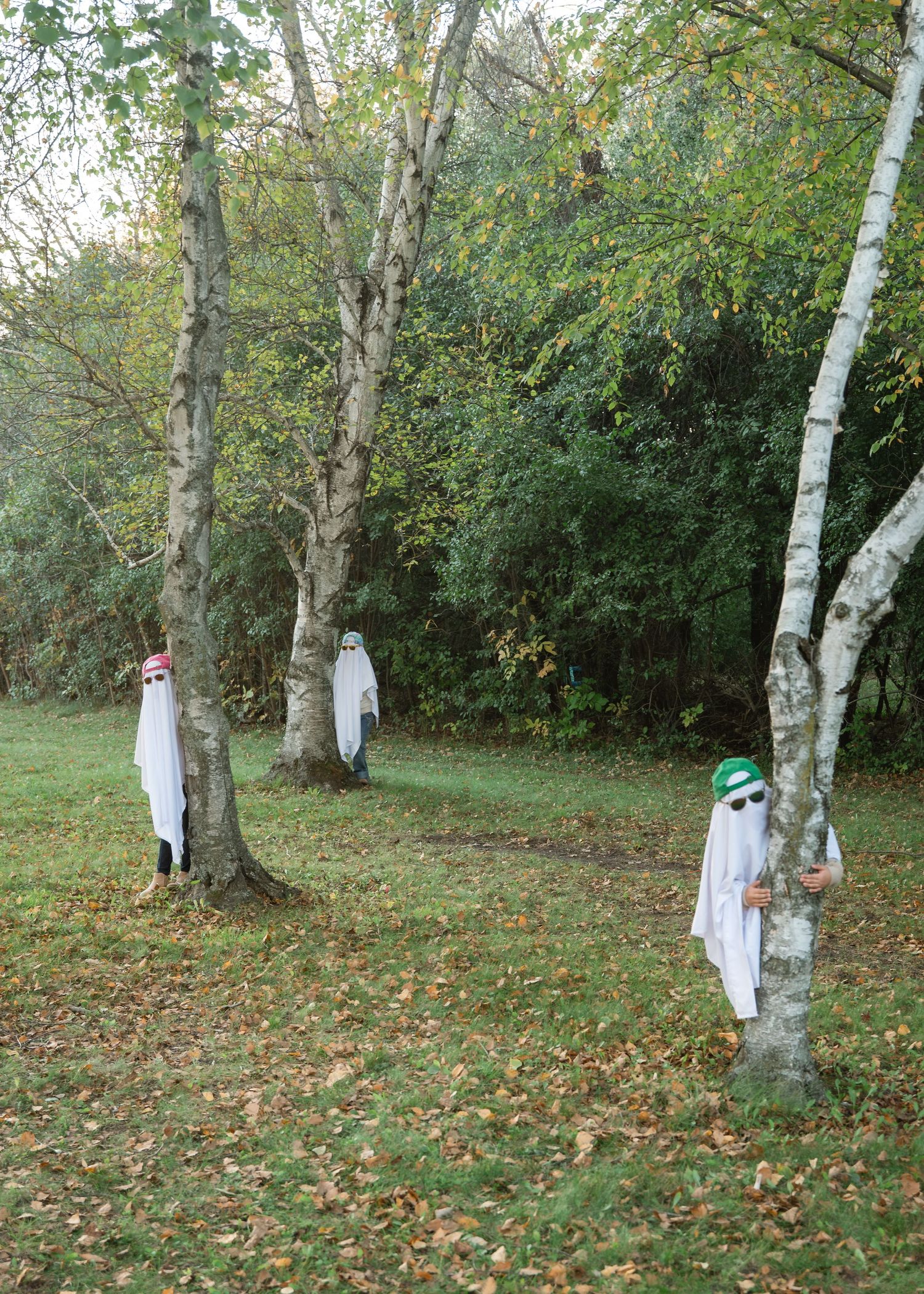 Three people in white sheet ghost costumes and colorful hats stand beside birch trees on a leaf-covered grassy lawn in autumn.