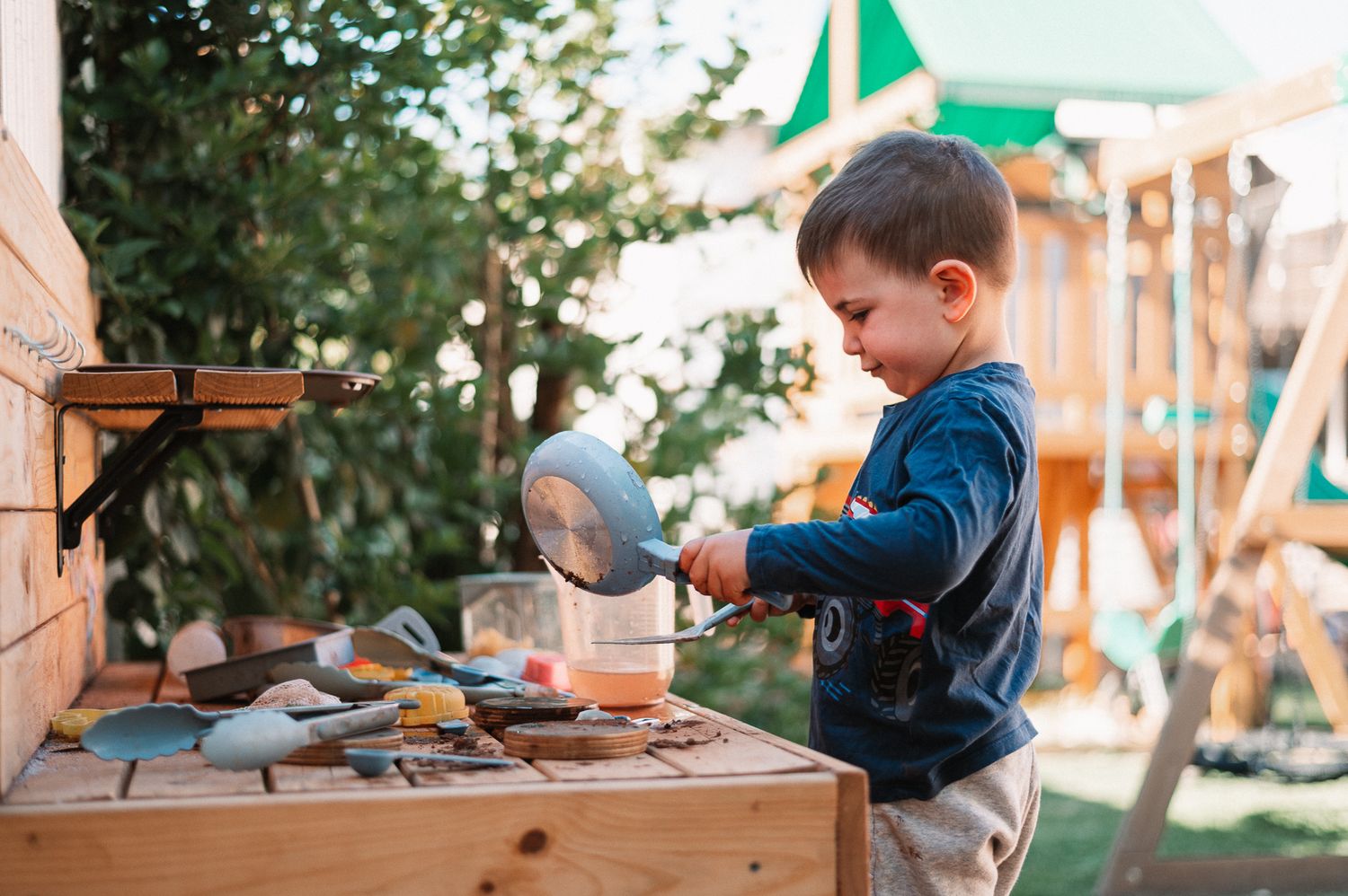 Toddler in a navy sweater plays with a metal pan and utensils at a wooden outdoor mud kitchen in a garden with a playground behind.
