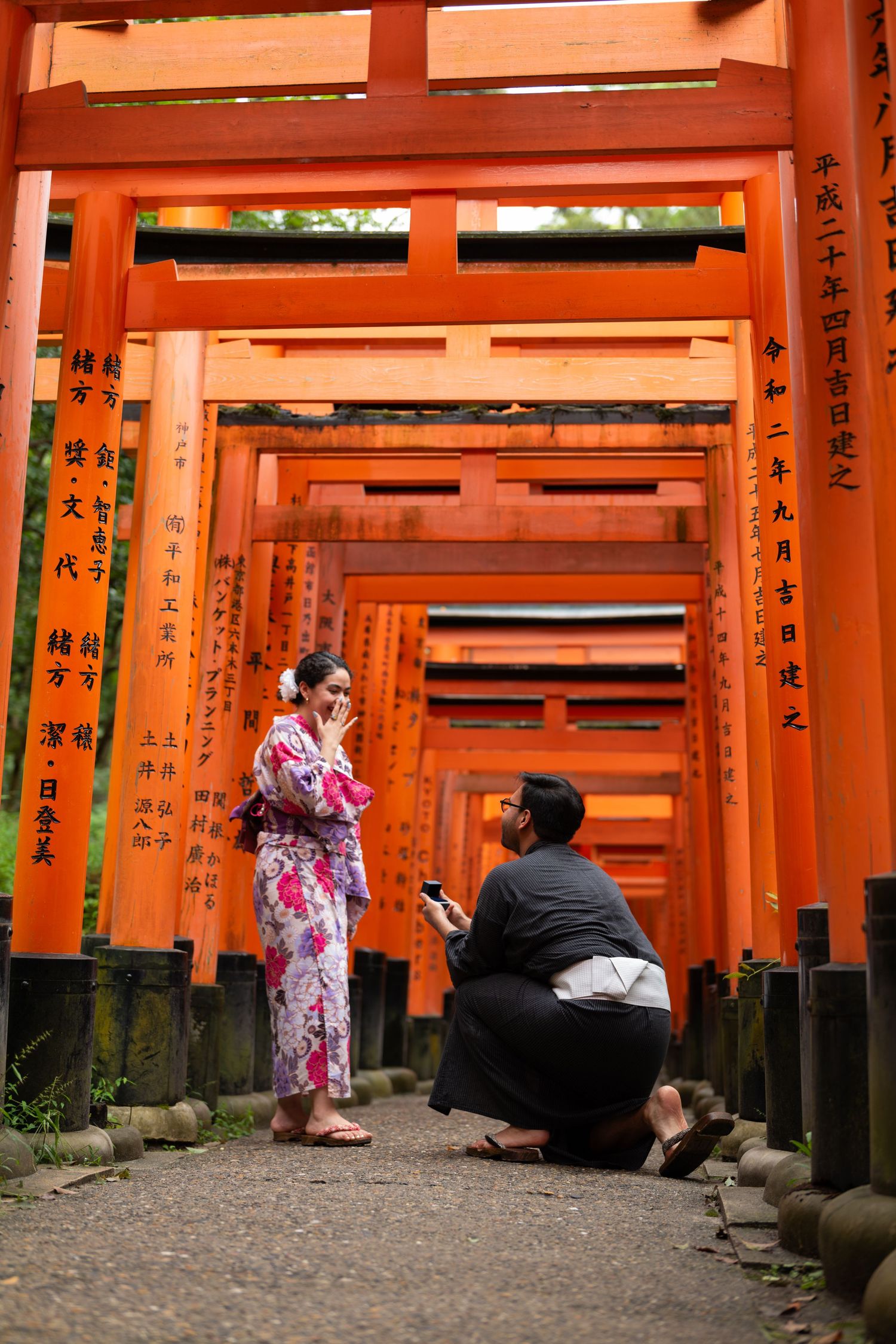 Man kneeling to propose to a smiling woman in a floral kimono among the vermilion torii gates of Fushimi Inari Shrine in Kyoto, Japan.