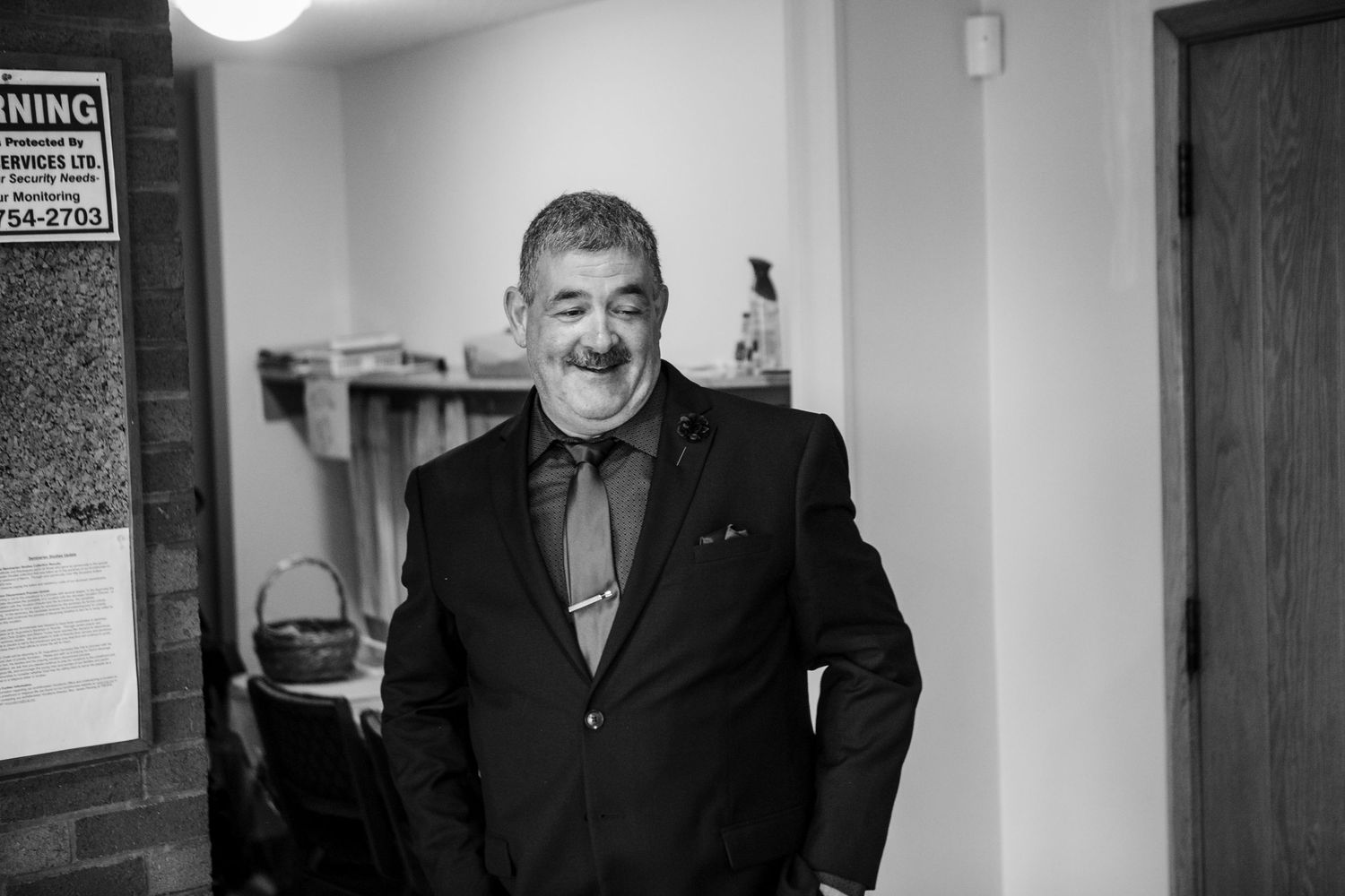 Smiling middle-aged man with a mustache wearing a dark suit and loosened tie stands in a small room in this black and white portrait.