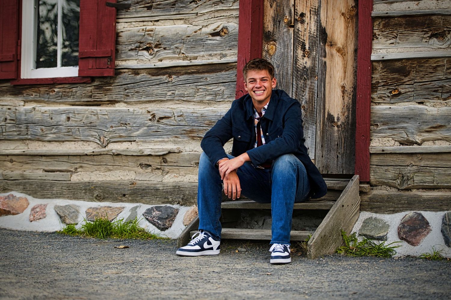 Smiling young man in a navy jacket, jeans and black-and-white sneakers sits on wooden steps in front of a rustic log cabin wall.