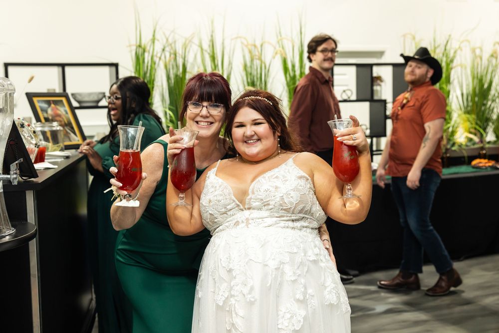 Two smiling women in a white lace dress and green dress raise red drinks at a festive gathering while guests mingle behind them.
