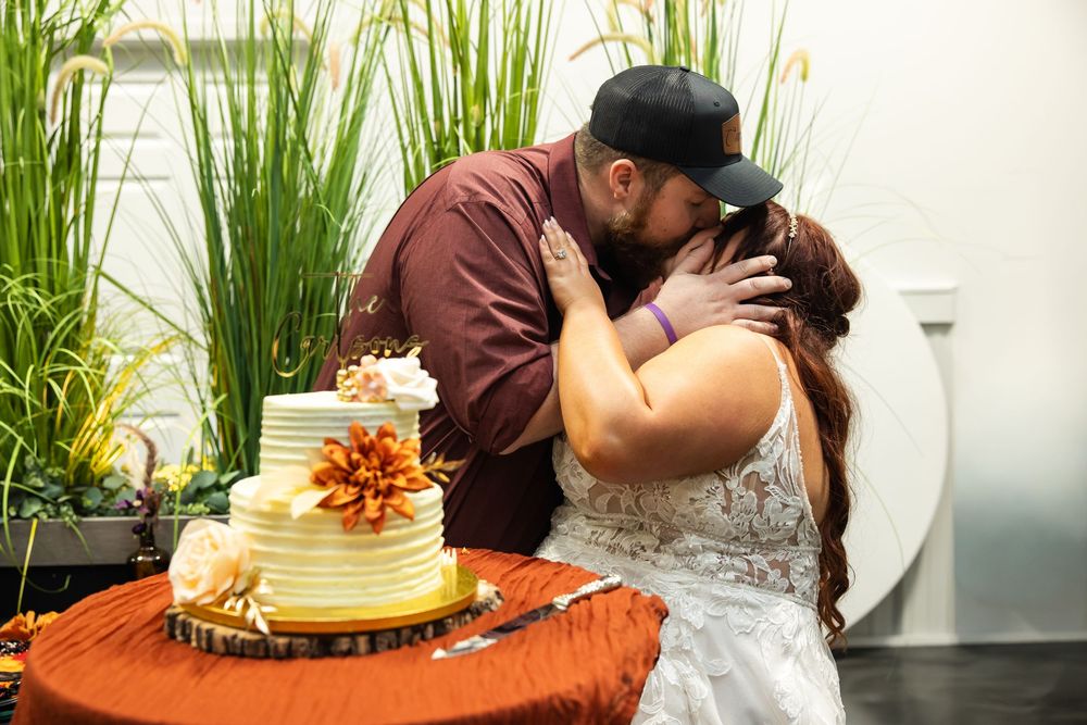 Bride and groom share a kiss beside a rustic wedding cake with orange flowers on a wood slice stand and orange table linen.