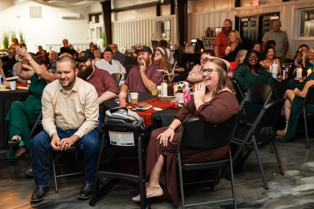Audience members laugh and smile while seated at tables during a lively indoor event in a rustic venue.