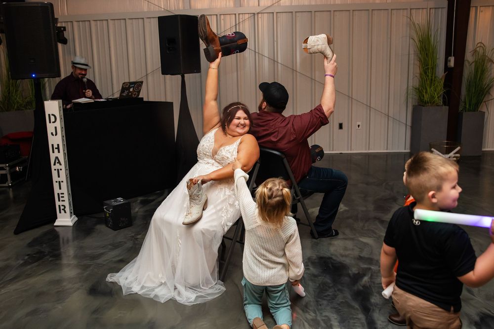 Bride in white gown sits in a chair holding up a garter as a man tosses it and children watch at a wedding reception with a DJ nearby.
