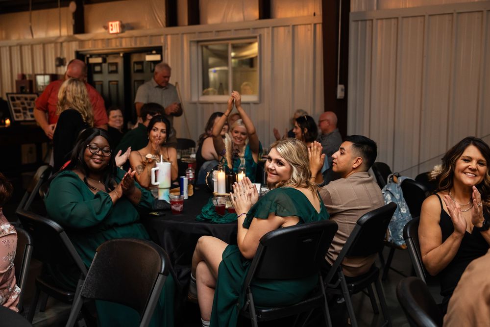 Guests in green dresses and casual attire applaud and smile at candlelit tables during an evening event.