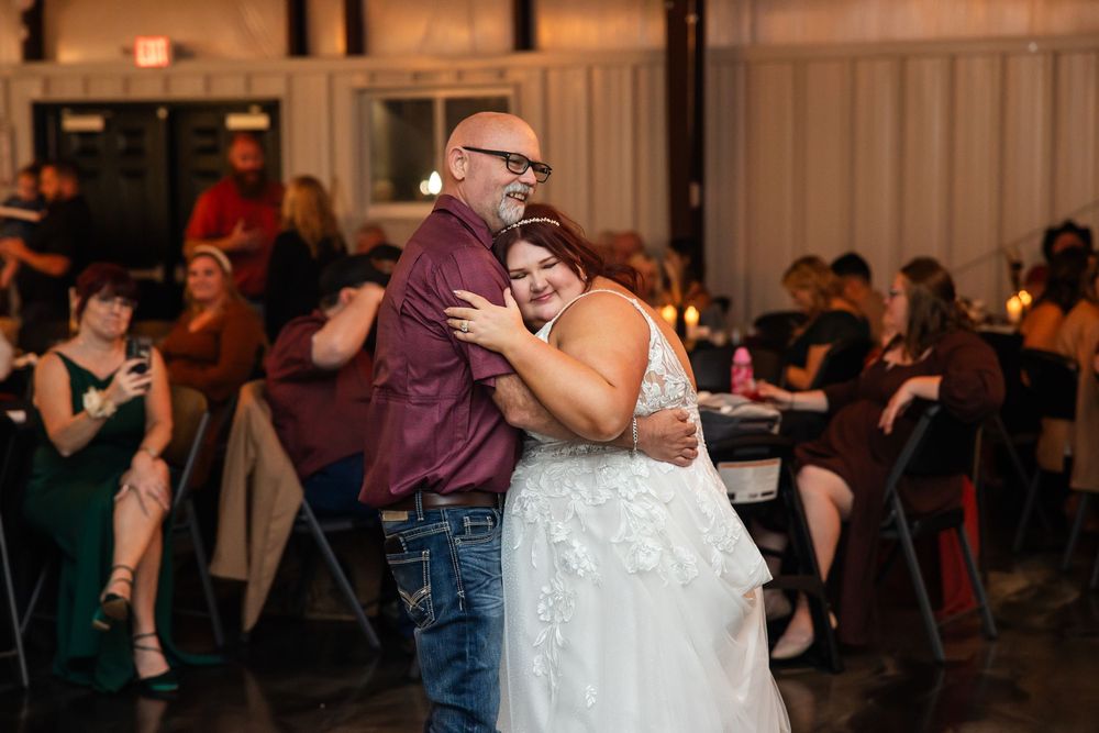 Bride in a white lace wedding dress embraces a bald, bearded man in a maroon shirt during a dance at a rustic wedding reception.