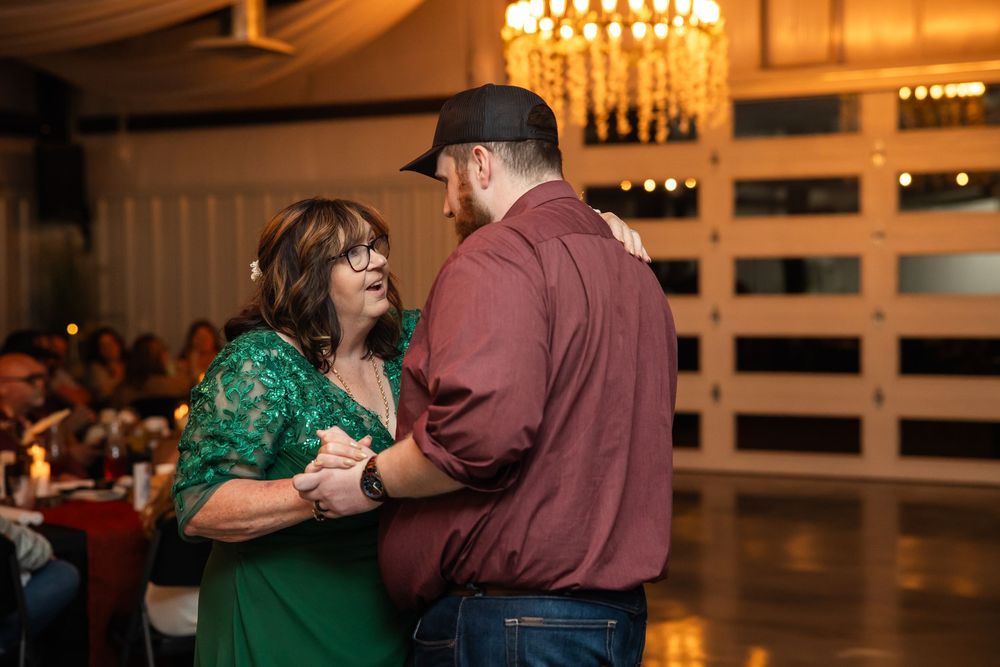 A woman in a green lace dress and a man in a maroon shirt and cap share a slow dance at a reception under a chandelier.