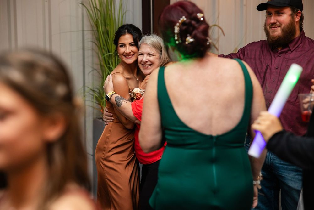 Two smiling women in dresses embrace at a lively party while a woman in a green dress faces them and a bearded man in a cap looks on.